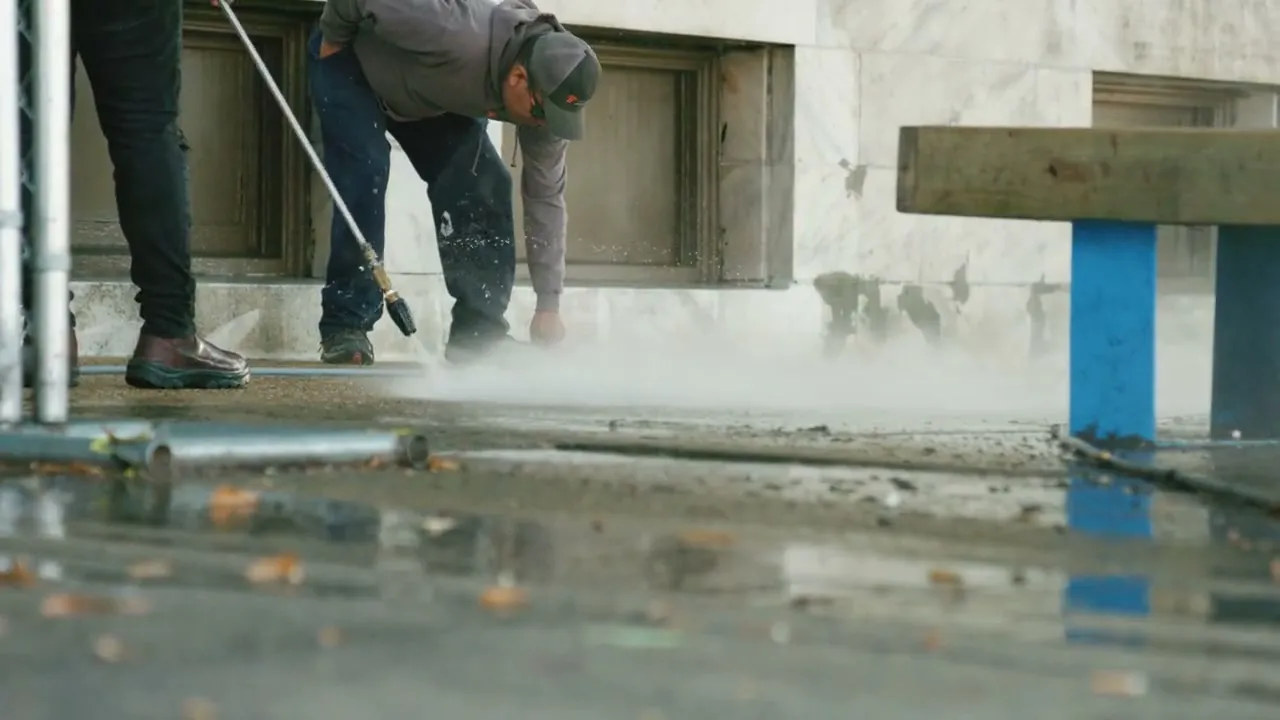 Staff member cleaning a concrete outdoor floor with the pressure washer, water spray visible
