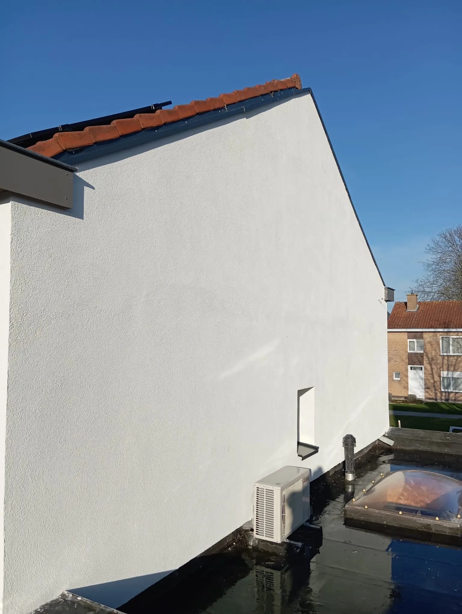 Strak afgewerkte witte crepi-gevel bij blauwe lucht, met plat dak op de voorgrond